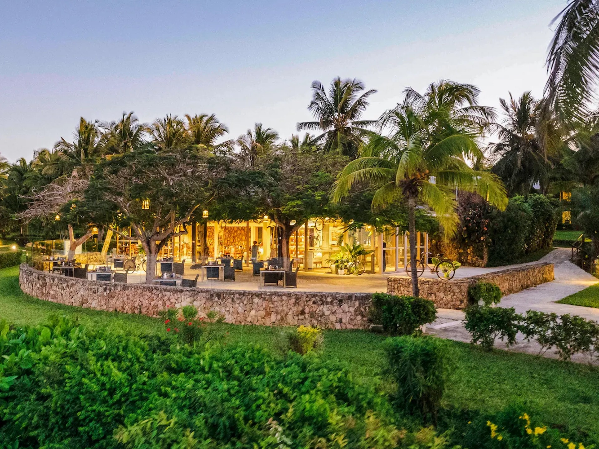A lamplit open-sided restaurant behind a coral-stone wall, set among coconut palms at dusk on the Tanzanian coast